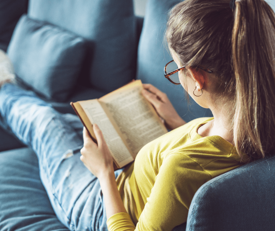 Brown haired girl laying on a blue couch reading a book. 